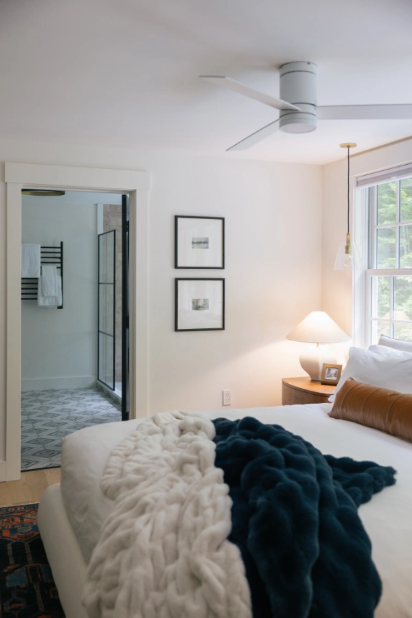 Primary bedroom view showing plush white bedding, layered throws, bedside lighting, and open doorway into ensuite bath